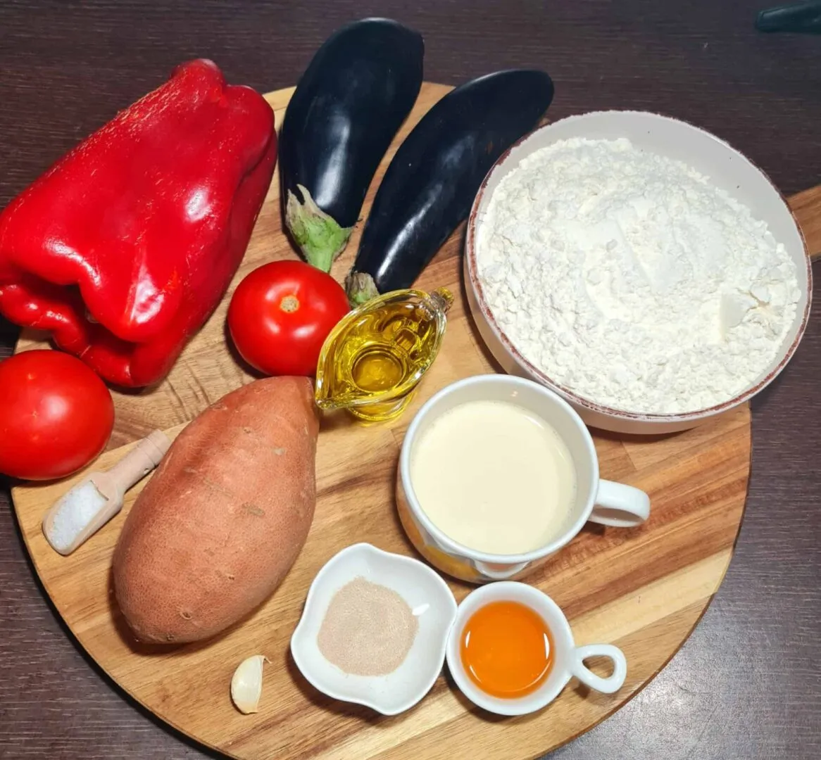 Sweet potato, red bell pepper, tomatoes, eggplants, flour, olive oil, yeast, garlic, salt, and plant milk arranged on a round wooden board for Sweet Potato Dinner Rolls.