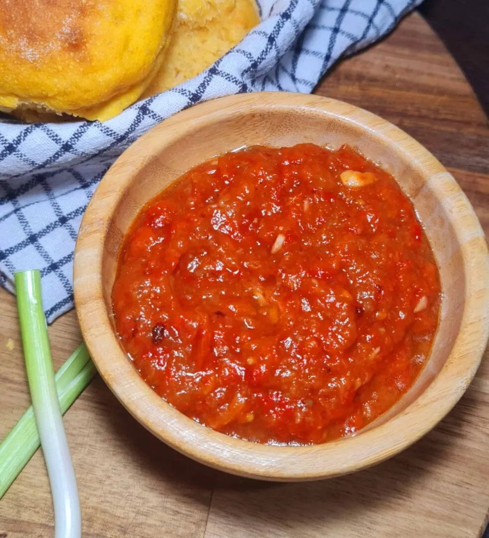 Wooden bowl filled with chunky roasted pepper sauce beside Sweet Potato Dinner Rolls wrapped in checkered cloth.
