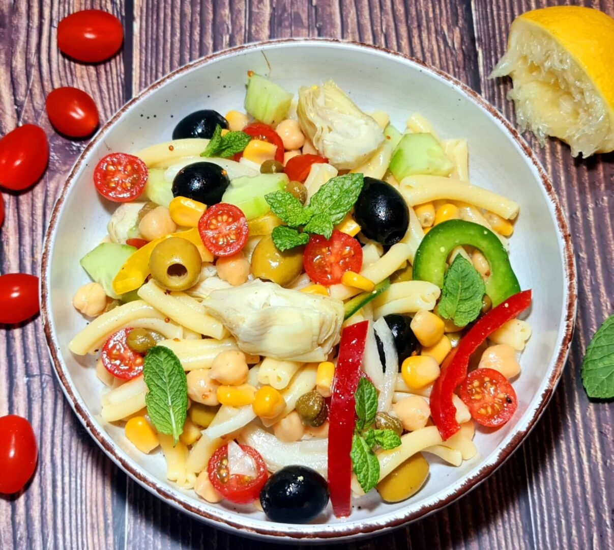Ceramic bowl of Mediterranean pasta salad with artichokes, olives, cucumber, tomato, corn, mint, and chickpeas on rustic wood.