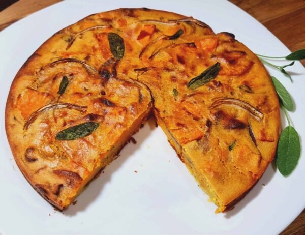 Whole sweet potato frittata on white plate, one slice cut out; crisp edges and sage leaves visible.