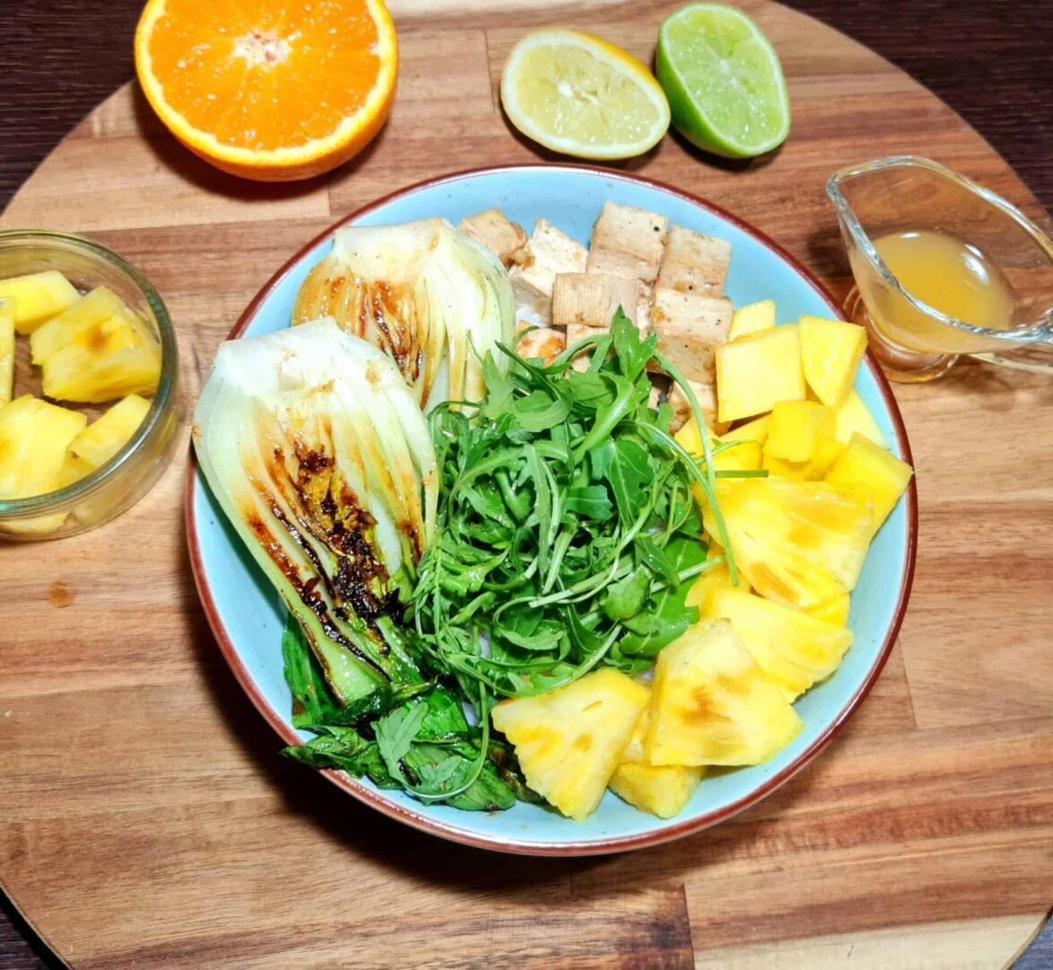 Top‑down shot of zesty vegan bowl featuring grilled bok choy, arugula, pineapple, tofu, and citrus wedges on a wooden board.