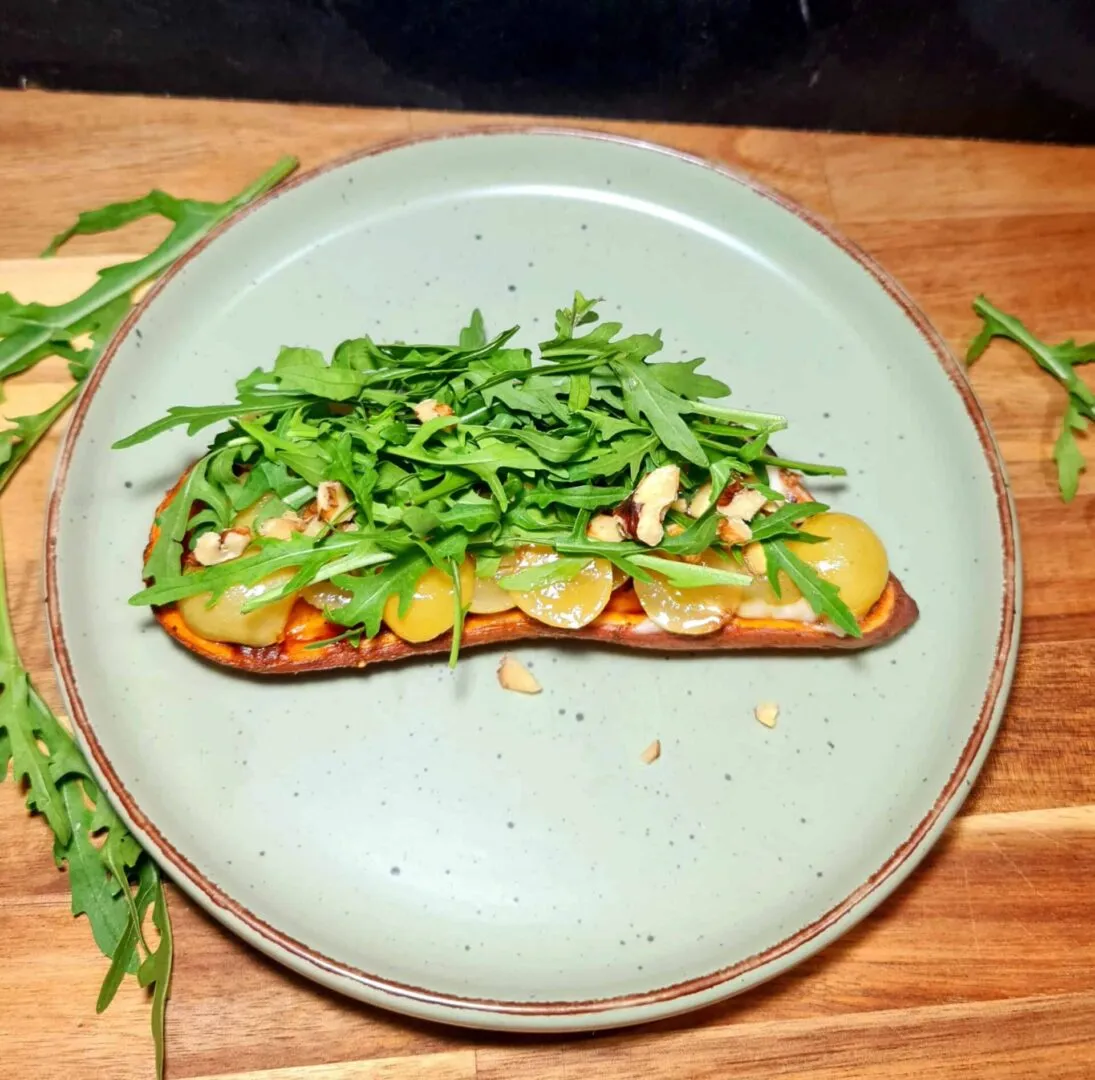 Overhead shot of sweet potato toast garnished with arugula, grapes and walnut pieces on pottery plate.
