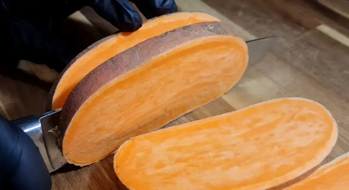Knife and gloved hand cutting long, flat slice from raw sweet potato on wooden board.