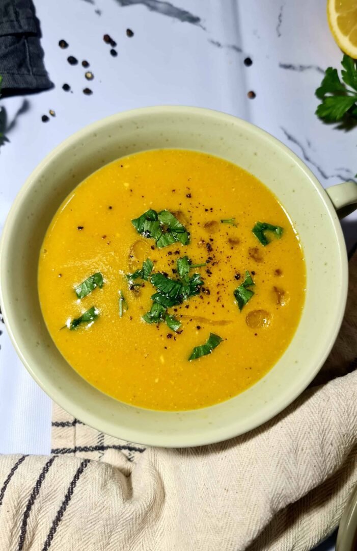 Close-up of a bowl of vibrant orange vegan lentil soup, garnished with fresh chopped parsley, black pepper, and a drizzle of olive oil.