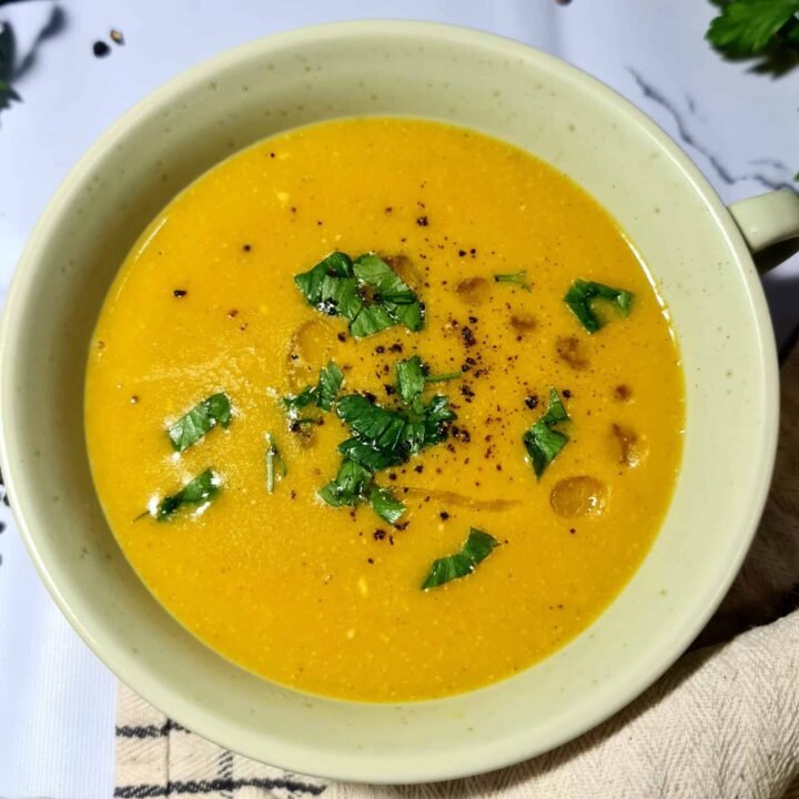 Close-up of a bowl of vibrant orange vegan lentil soup, garnished with fresh chopped parsley, black pepper, and a drizzle of olive oil.