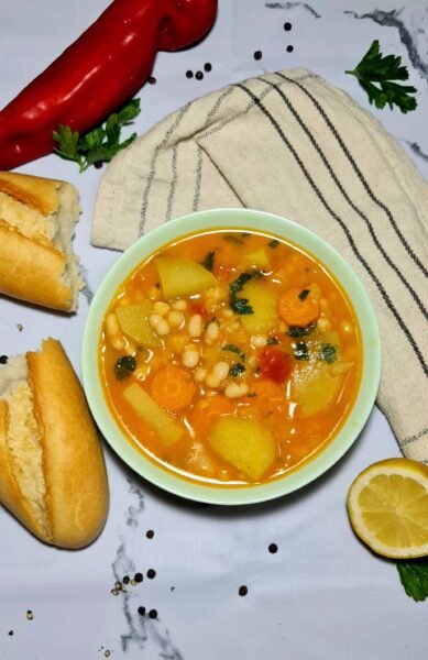 An overhead shot of a bowl of potato and white bean soup on a marble surface, surrounded by a torn baguette, a red pepper, a lemon slice, and a kitchen towel.