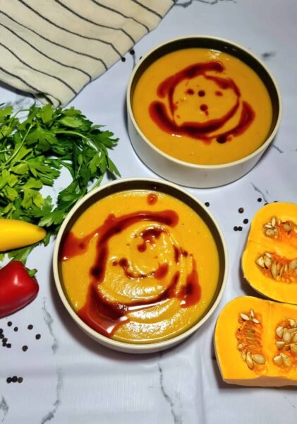 Two bowls of creamy pumpkin-lentil soup topped with chili oil swirl, surrounded by parsley, mini peppers, and squash halves.