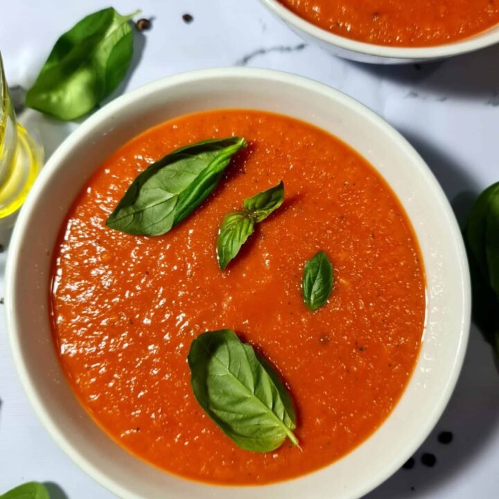 A close-up shot of a white bowl filled with vibrant red tomato basil soup, garnished with several fresh green basil leaves.