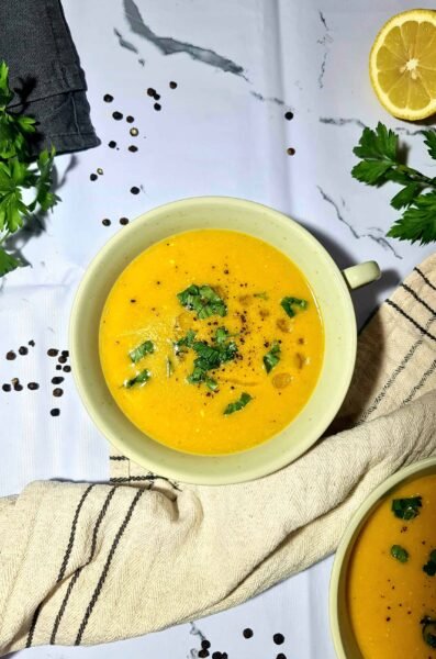 Overhead shot of two bowls of vibrant orange vegan lentil soup garnished with fresh parsley, black pepper, and olive oil, with a lemon half and peppercorns on a white marble background.