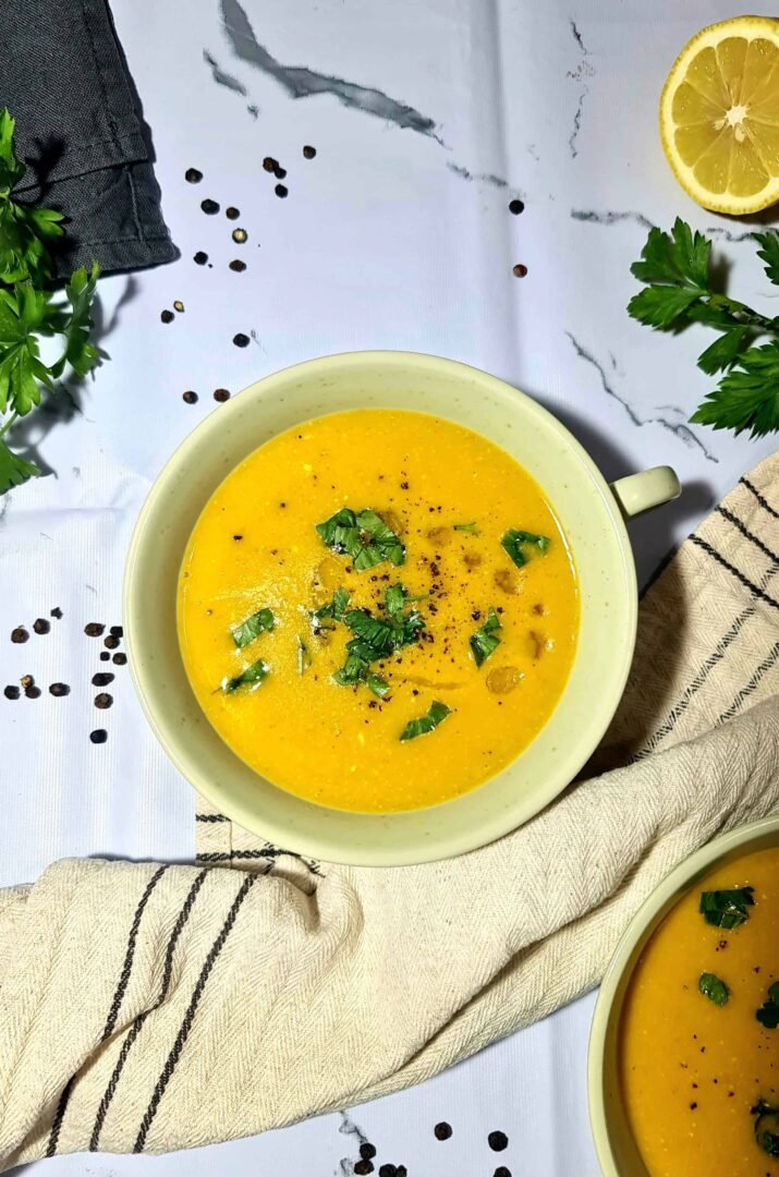 Overhead shot of two bowls of vibrant orange vegan lentil soup garnished with fresh parsley, black pepper, and olive oil, with a lemon half and peppercorns on a white marble background.