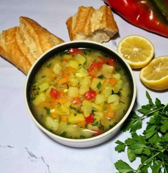 A black and white bowl of easy vegan potato soup garnished with parsley, served with lemon slices and pieces of crusty French bread on a white marble background.