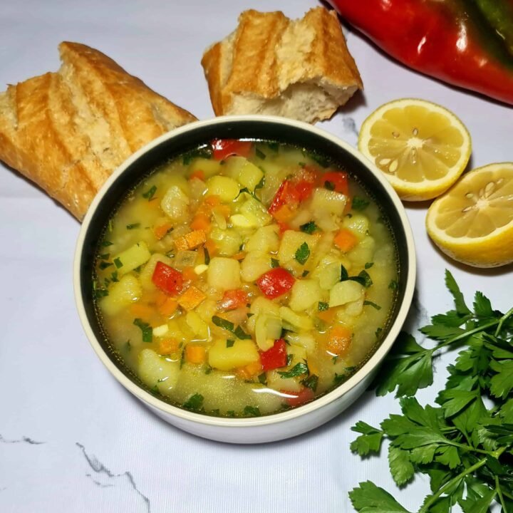 A black and white bowl of easy vegan potato soup garnished with parsley, served with lemon slices and pieces of crusty French bread on a white marble background.