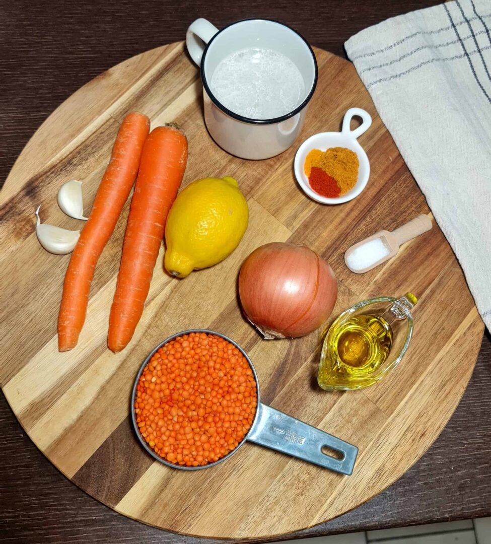 Overhead shot of vegan lentil soup ingredients arranged on a wooden board: red lentils, carrots, lemon, onion, garlic, vegetable milk, olive oil, salt, turmeric, and paprika.