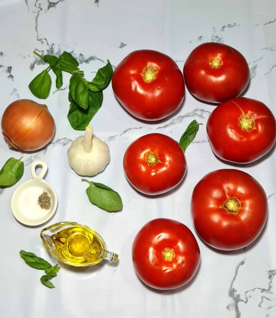 Fresh ingredients for tomato basil soup arranged on a white marble background: ripe tomatoes, a whole onion, a bulb of garlic, fresh basil leaves, olive oil, and seasoning.