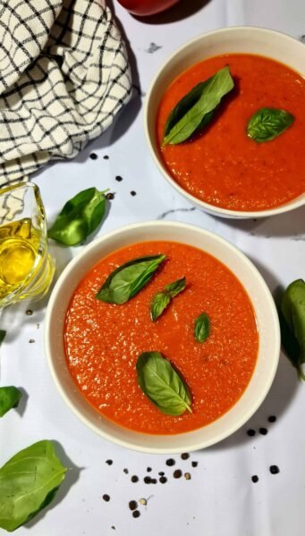 Two white bowls of homemade tomato basil soup garnished with fresh basil, with a small pitcher of olive oil and scattered peppercorns on a marble surface.