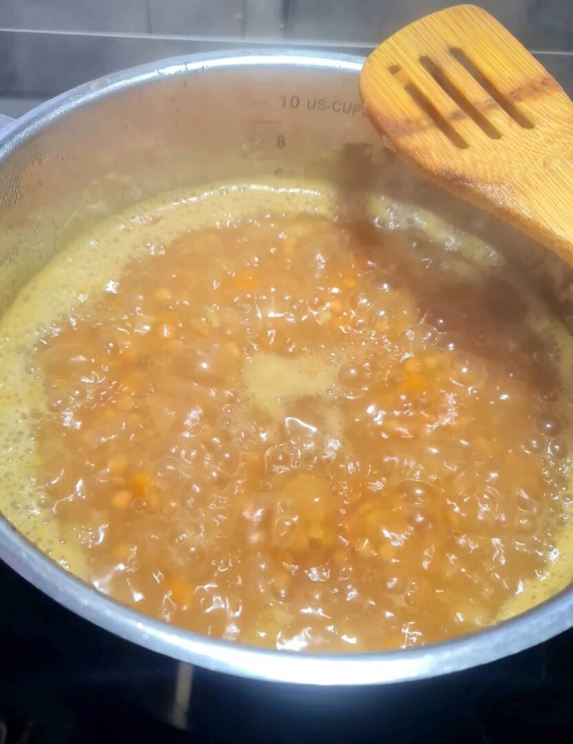 Red lentils and vegetables simmering in a pot of golden liquid on a stove, with steam rising and a wooden spoon resting on the edge.