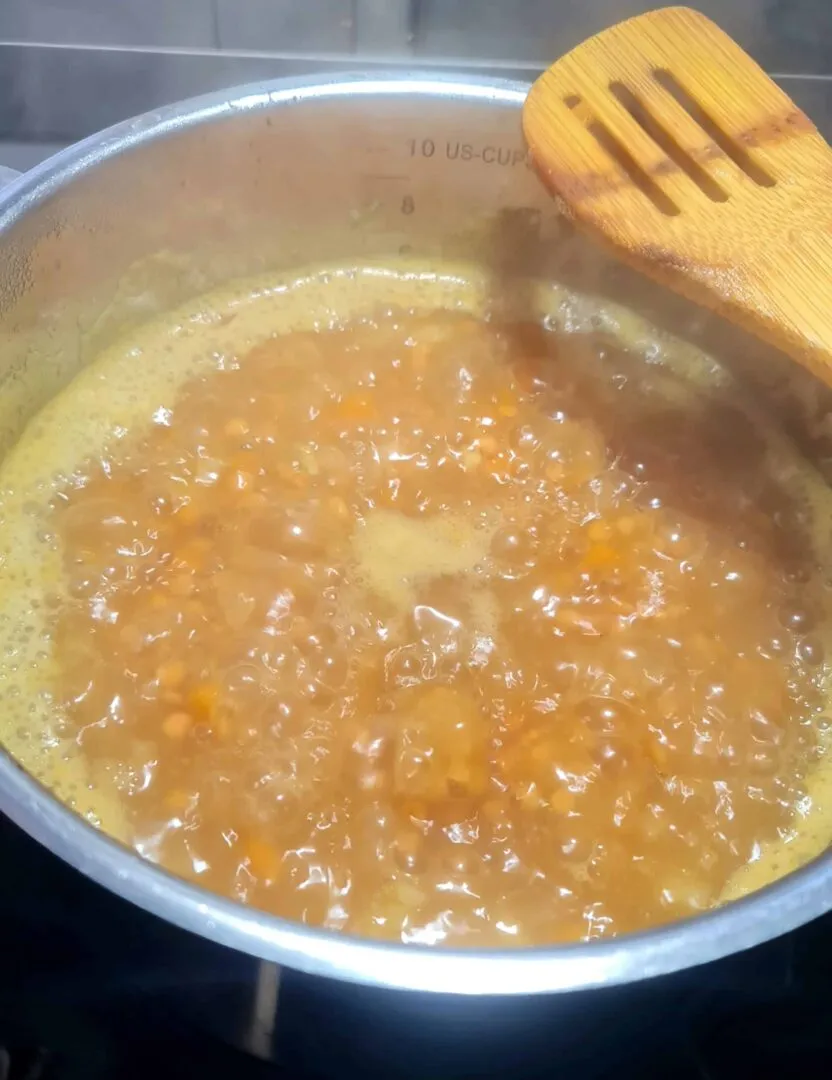 Red lentils and vegetables simmering in a pot of golden liquid on a stove, with steam rising and a wooden spoon resting on the edge.