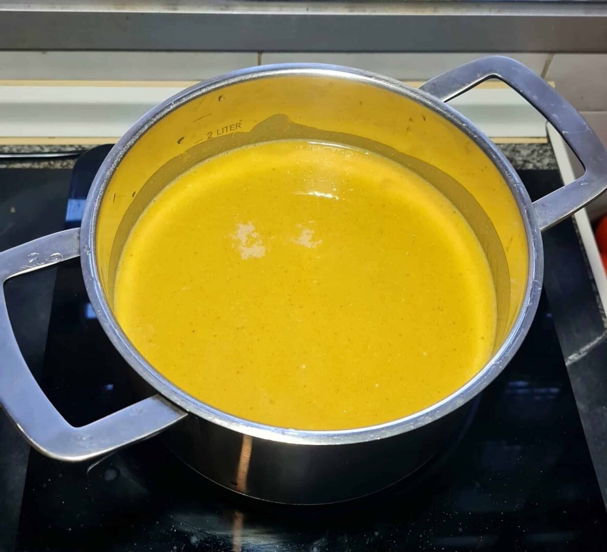 Smooth, vibrant orange vegan lentil soup resting in a stainless steel pot on an induction stovetop after blending, ready to serve.