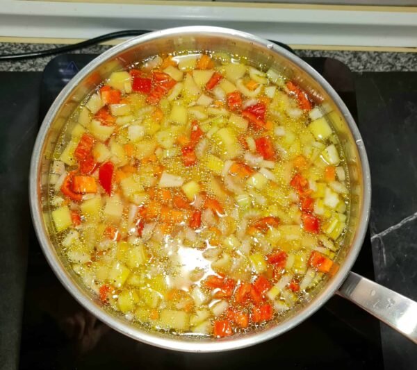 Diced potatoes, red peppers, and onions simmering in a clear vegetable broth inside a silver pot on a black stovetop, a step in the easy vegan potato soup recipe.