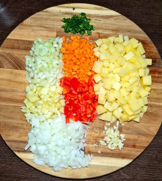 Diced potatoes, carrots, onions, celery, and red peppers arranged on a round wooden cutting board, ready to make easy vegan potato soup.
