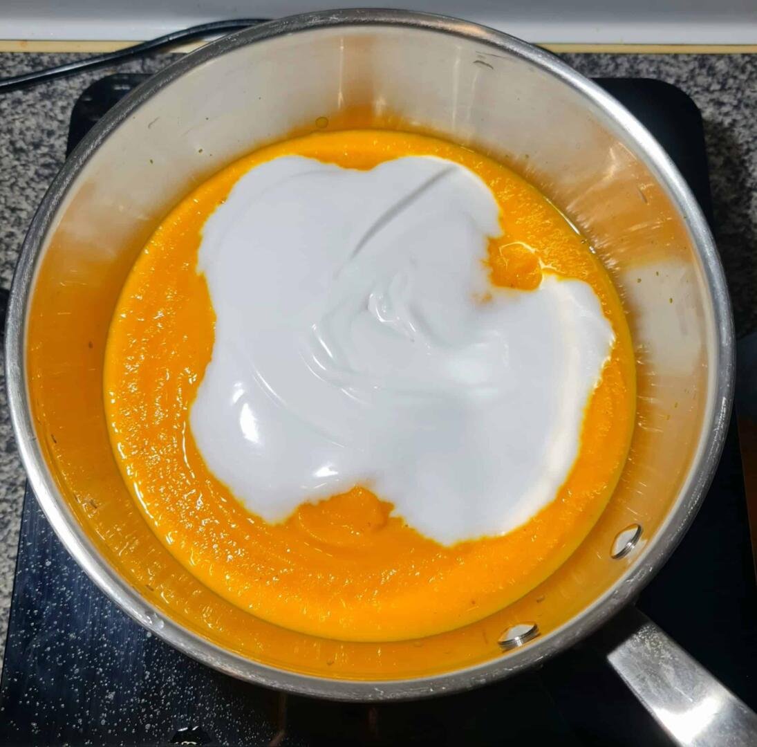 Thick, white coconut milk being poured into a vibrant orange pureed carrot soup in a stainless steel pot.