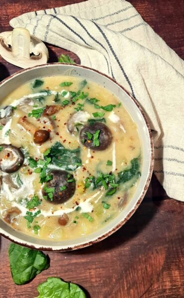 A close-up, angled shot of a bowl of creamy potato mushroom soup, showing chunks of mushrooms and spinach, and garnished with fresh parsley, red pepper flakes, and a swirl of vegan cream.