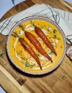 A rustic bowl of creamy orange roasted carrot potato soup, elegantly garnished with three whole roasted carrots, a swirl of coconut cream, and fresh parsley, set on a wooden board.