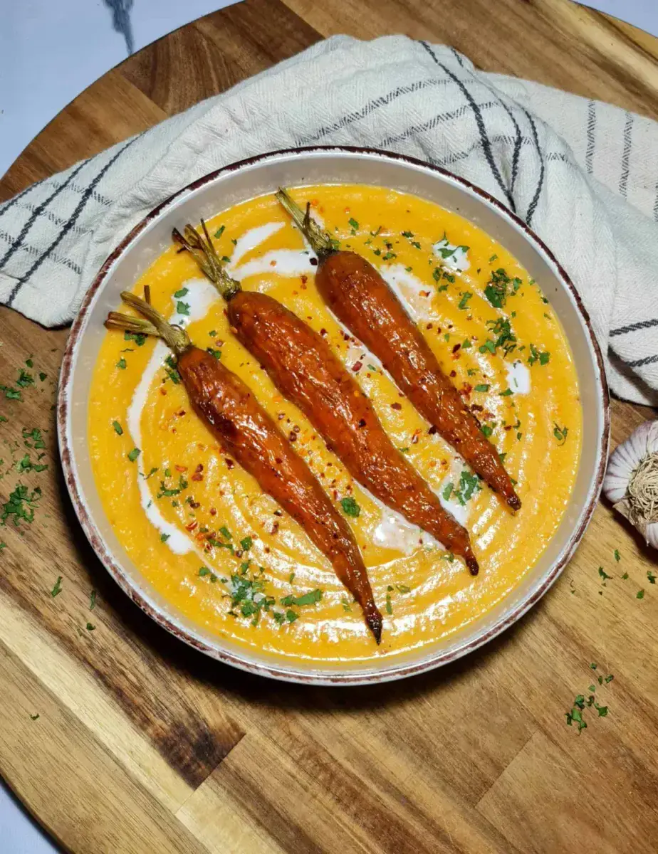 A rustic bowl of creamy orange roasted carrot potato soup, elegantly garnished with three whole roasted carrots, a swirl of coconut cream, and fresh parsley, set on a wooden board.