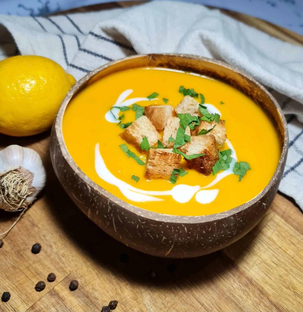 A rustic coconut bowl filled with bright orange carrot ginger soup, garnished with crispy croutons, fresh parsley, and a swirl of coconut cream, set on a wooden board.