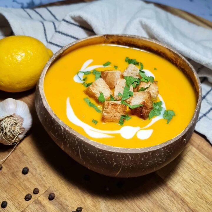 A rustic coconut bowl filled with bright orange carrot ginger soup, garnished with crispy croutons, fresh parsley, and a swirl of coconut cream, set on a wooden board.