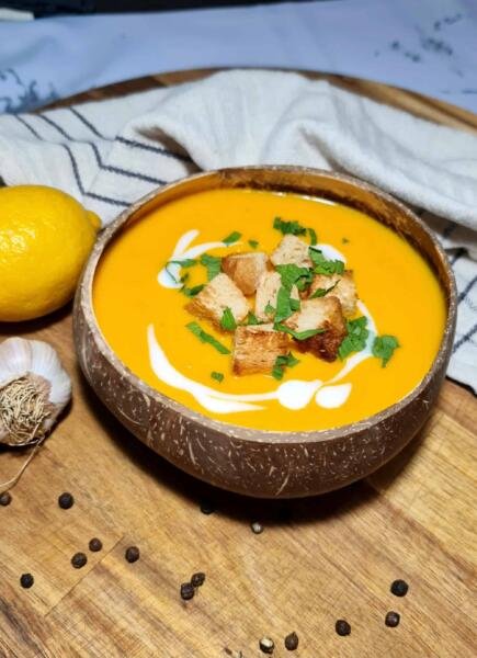 A rustic coconut bowl filled with bright orange carrot ginger soup, garnished with crispy croutons, fresh parsley, and a swirl of coconut cream, set on a wooden board.