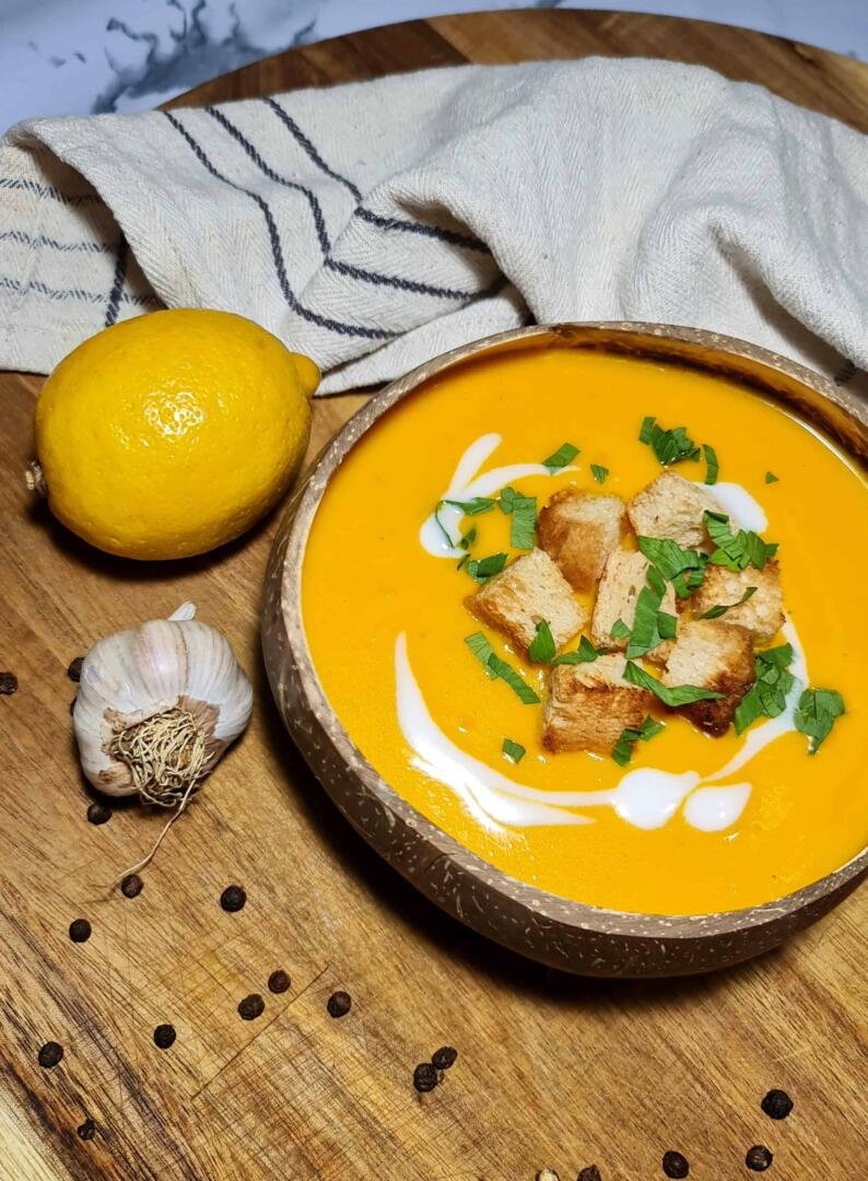 A rustic coconut bowl filled with bright orange carrot ginger soup, garnished with crispy croutons, fresh parsley, and a swirl of coconut cream, set on a wooden board.