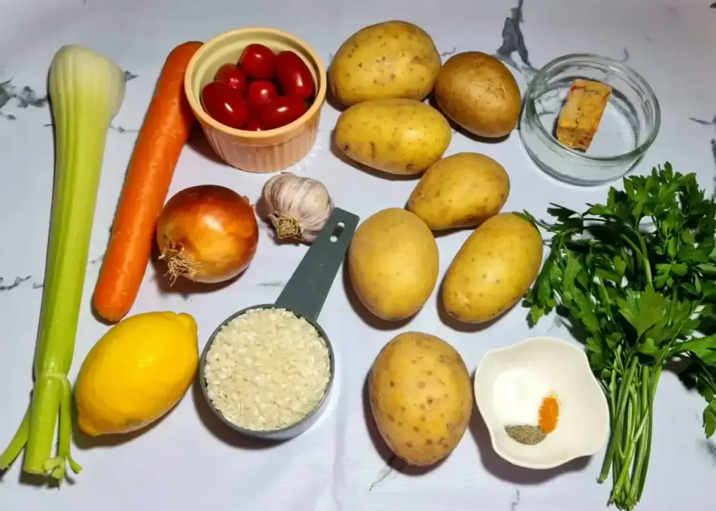 Ingredients for a vegan potato rice soup on a marble surface: whole potatoes, a carrot, celery, onion, garlic, rice, cherry tomatoes, lemon, and fresh parsley.