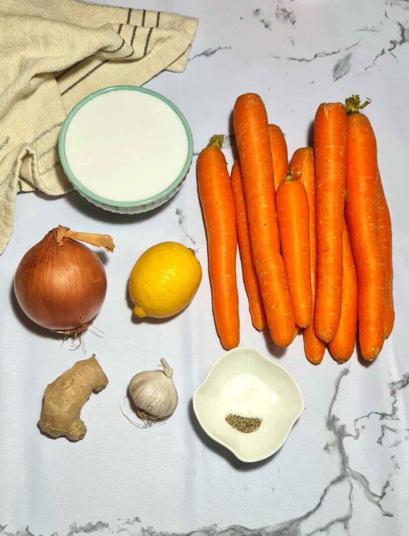 Ingredients for a vegan carrot ginger soup on a marble background: a bunch of fresh carrots, an onion, a knob of ginger, a lemon, garlic, a bowl of coconut milk, and seasoning.