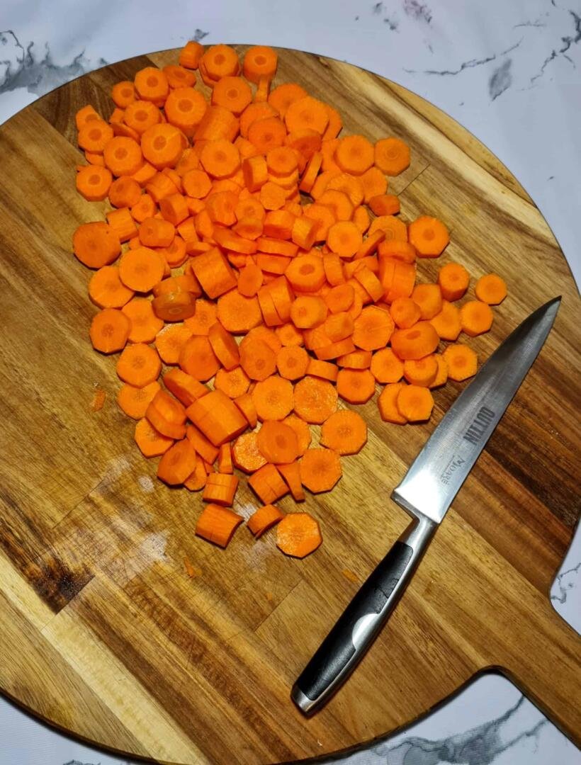 A large pile of bright orange sliced carrots on a round wooden cutting board with a sharp kitchen knife resting beside it.