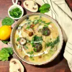 An overhead shot of a rustic bowl of creamy potato mushroom soup, styled on a dark wood table with a fresh lemon, spinach leaves, chopped parsley, and a kitchen towel. vegan fall and winter soup recipes