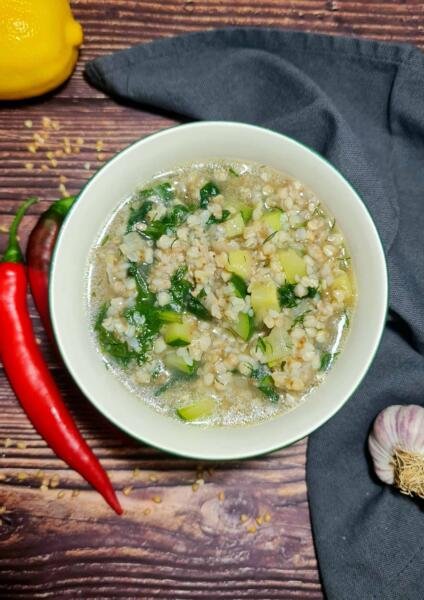 A white bowl filled with nourishing buckwheat soup, showing cooked buckwheat groats, chunks of zucchini, and fresh spinach in a clear broth, styled on a dark wood table with fresh chili and lemon.(1)