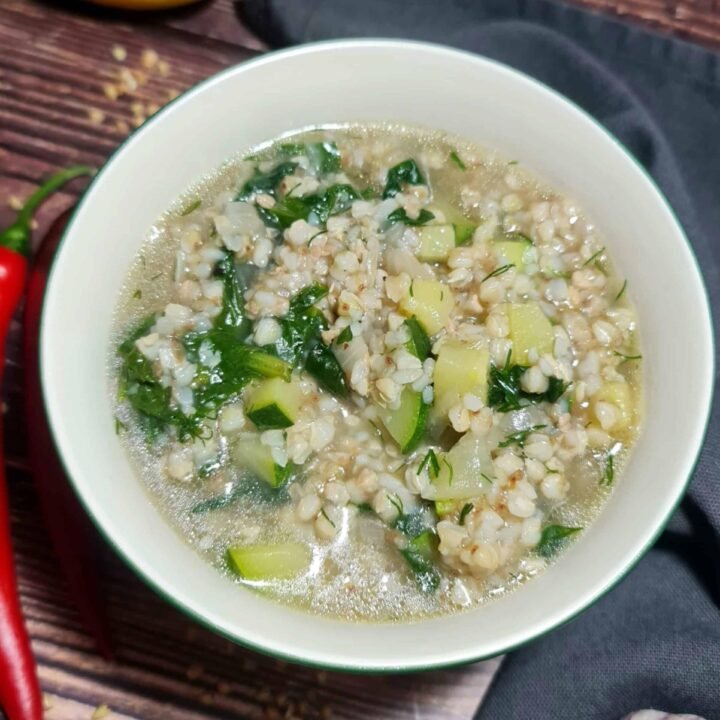 A white bowl filled with nourishing buckwheat soup, showing cooked buckwheat groats, chunks of zucchini, and fresh spinach in a clear broth, styled on a dark wood table with fresh chili and lemon.