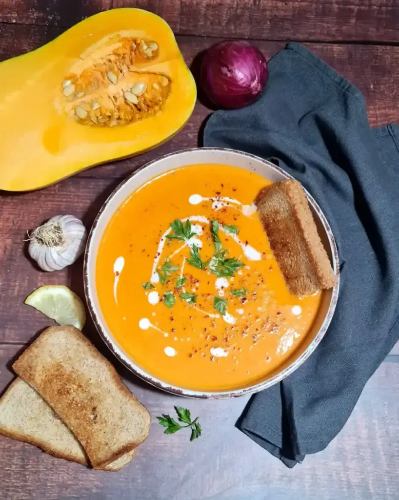A rustic bowl of creamy roasted winter vegetable soup, garnished with fresh parsley, coconut cream, and chili flakes, served with toasted bread on a dark wood table.