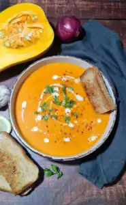 A rustic bowl of creamy roasted winter vegetable soup, garnished with fresh parsley, coconut cream, and chili flakes, served with toasted bread on a dark wood table.