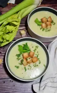Two rustic bowls of creamy, light green potato celery soup, garnished with golden-brown croutons and fresh parsley, set on a dark wood table.