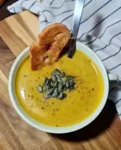 Creamy pumpkin zucchini soup topped with pumpkin seeds, served with toasted bread on a wooden board.