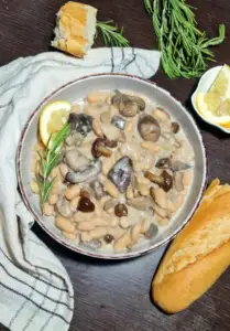 Top-down view of a rustic bowl filled with creamy mushroom and white beans, served with a piece of crusty bread, fresh rosemary sprigs, and lemon wedges on a dark wooden background.