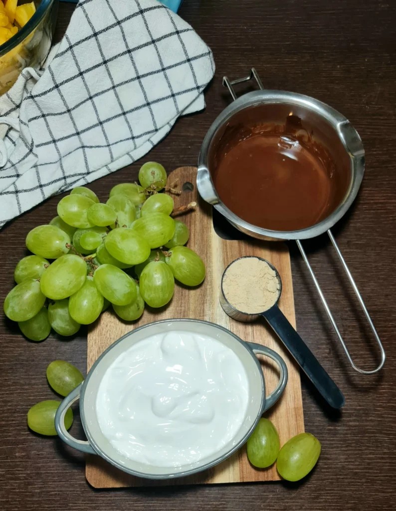 Fresh green grapes, a bowl of yogurt, protein powder, and melted chocolate set out to make high protein chocolate yogurt grapes.