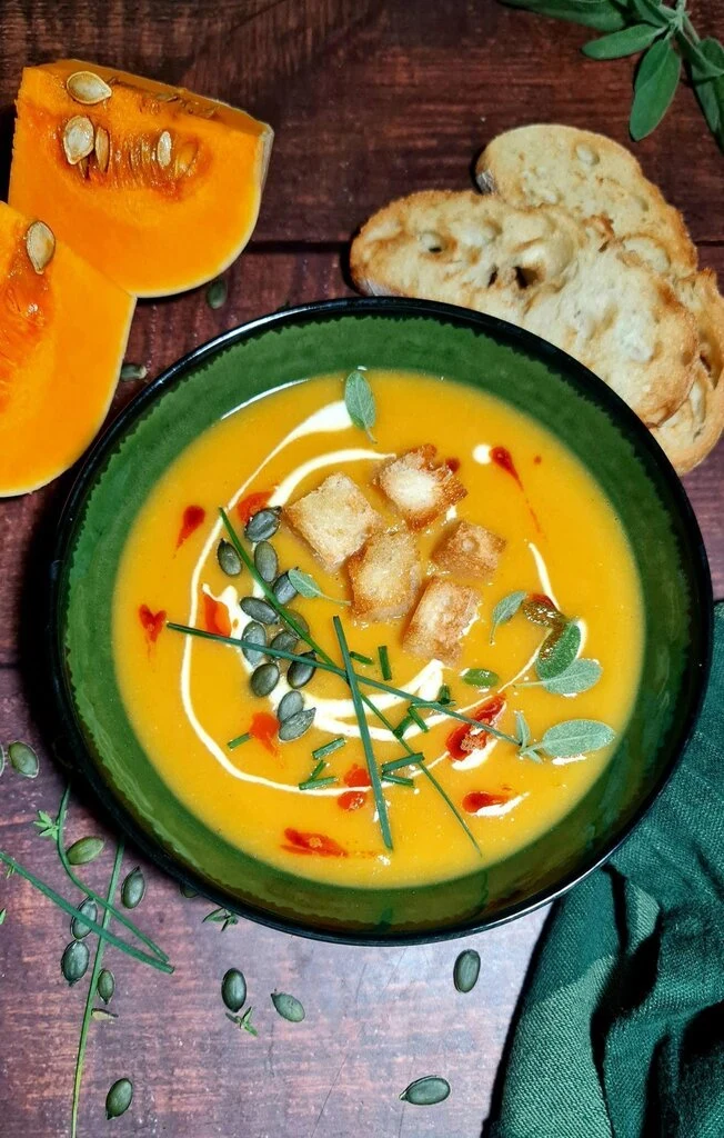 Finished creamy vegan pumpkin soup in green bowl garnished with crispy croutons, toasted pumpkin seeds, fresh sage and chives, cream swirl, and chili oil, served with fresh pumpkin wedges and crusty bread