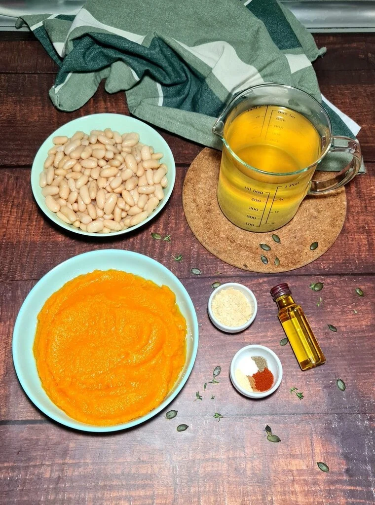 Ingredients for creamy vegan pumpkin soup on rustic wooden table: pumpkin puree, cannellini beans, vegetable broth in measuring cup, nutritional yeast, olive oil, and spices with green linen napkin