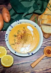 Top-down view of creamy sweet potato and lentil soup in a bowl with a coconut cream swirl, fresh thyme, and red pepper flakes, accompanied by lemon halves, sliced bread, and raw sweet potatoes.