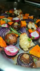 Angled view of a metal baking tray filled with golden-brown sheet pan fall veggies, featuring crispy roasted cauliflower florets, root vegetables, and fresh rosemary sprigs.