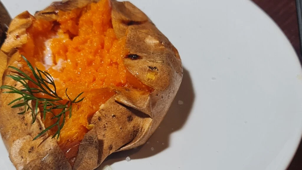 A close-up view of a freshly baked sweet potato with melting butter and a sprig of fresh green dill on a white plate. Learn how to bake sweet potatoes.