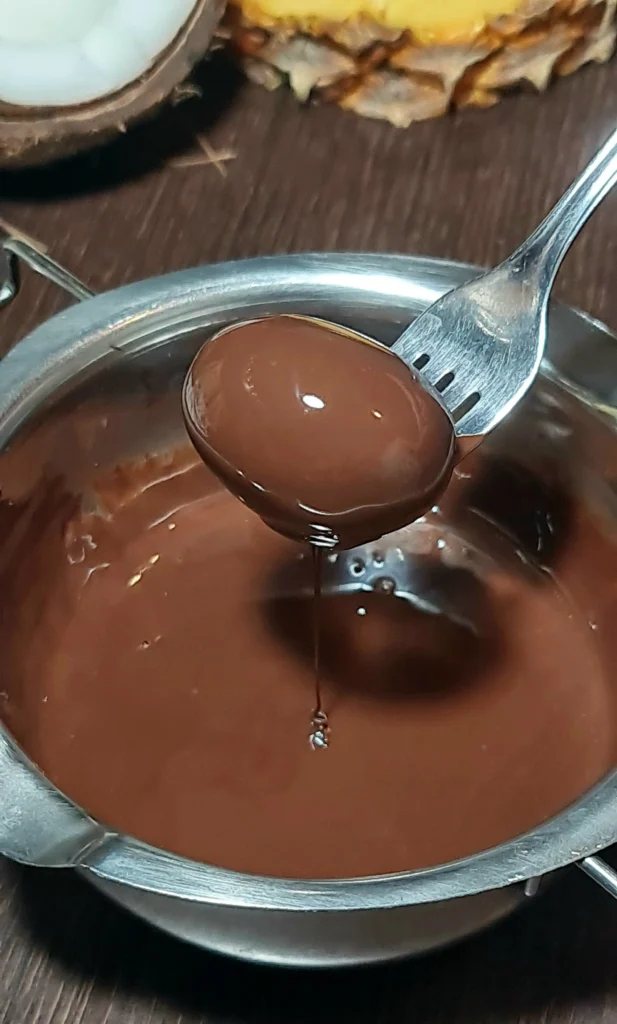 A frozen, yogurt-coated grape held by a fork being dipped into a pot of melted chocolate to create chocolate grape bark